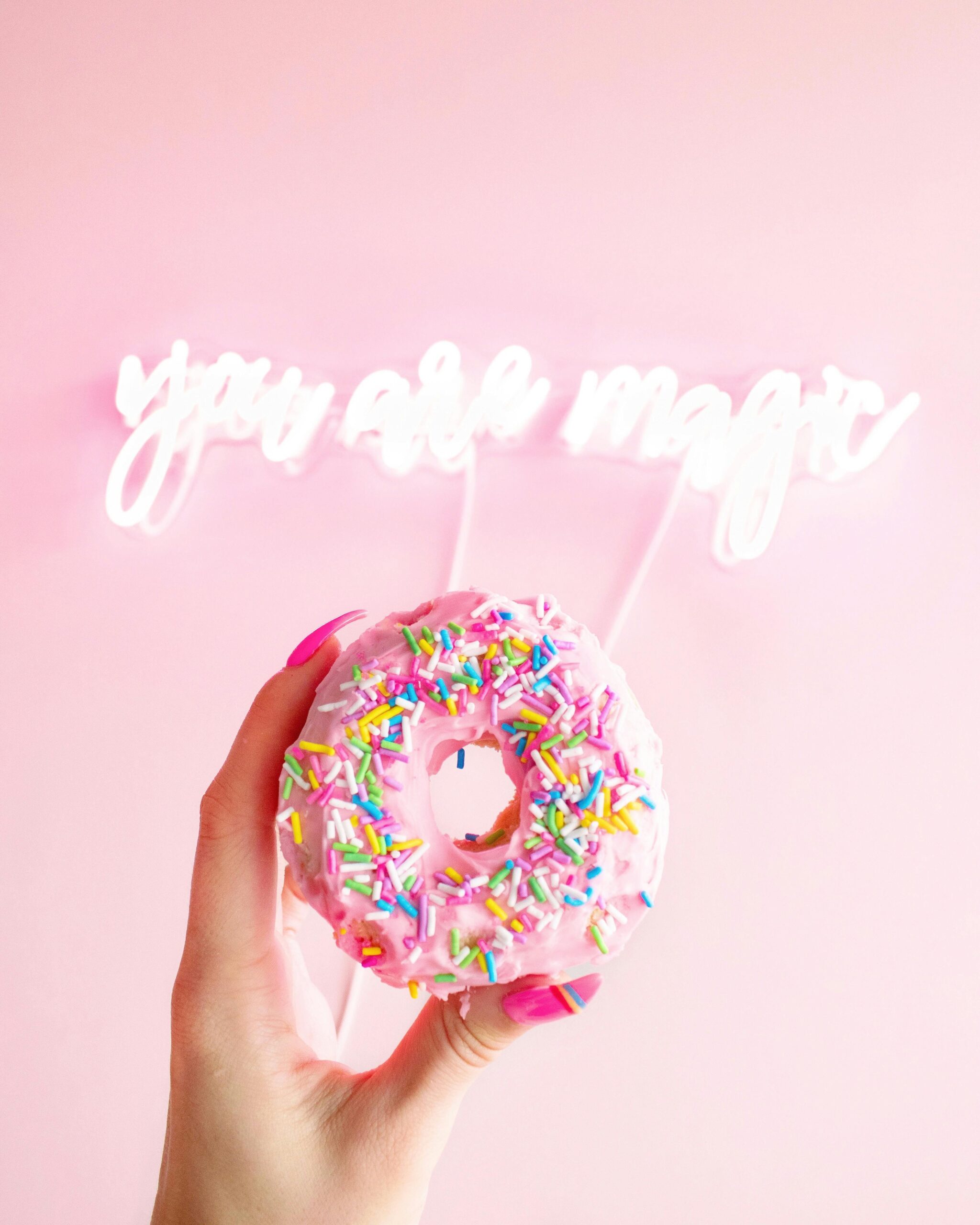 Behandelingen & tarieven Vibrant donut with sprinkles held against a pink background and neon 'you are magic' sign.